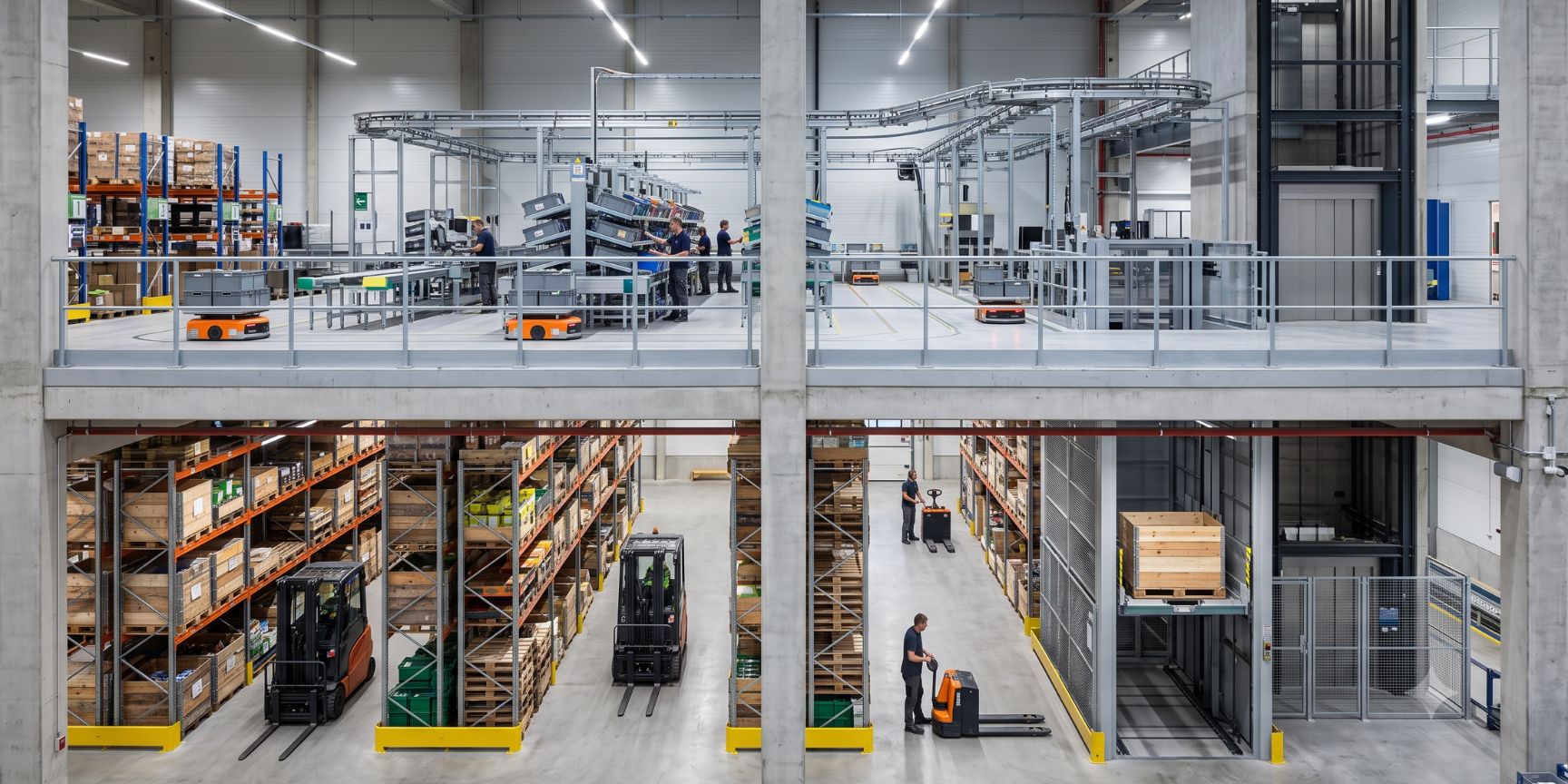Interior view of multi-storey logistics hall: ground floor with forklift trucks, upper floor with automated e-commerce picking and conveyor technology. Networked intralogistics.